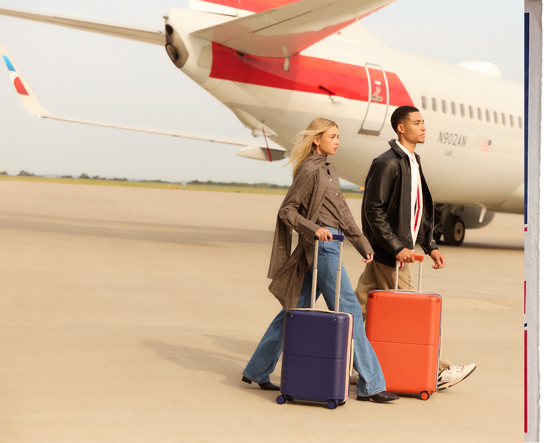 Two travellers walking a tarmac beside an American Airlines aircraft with July suitcases