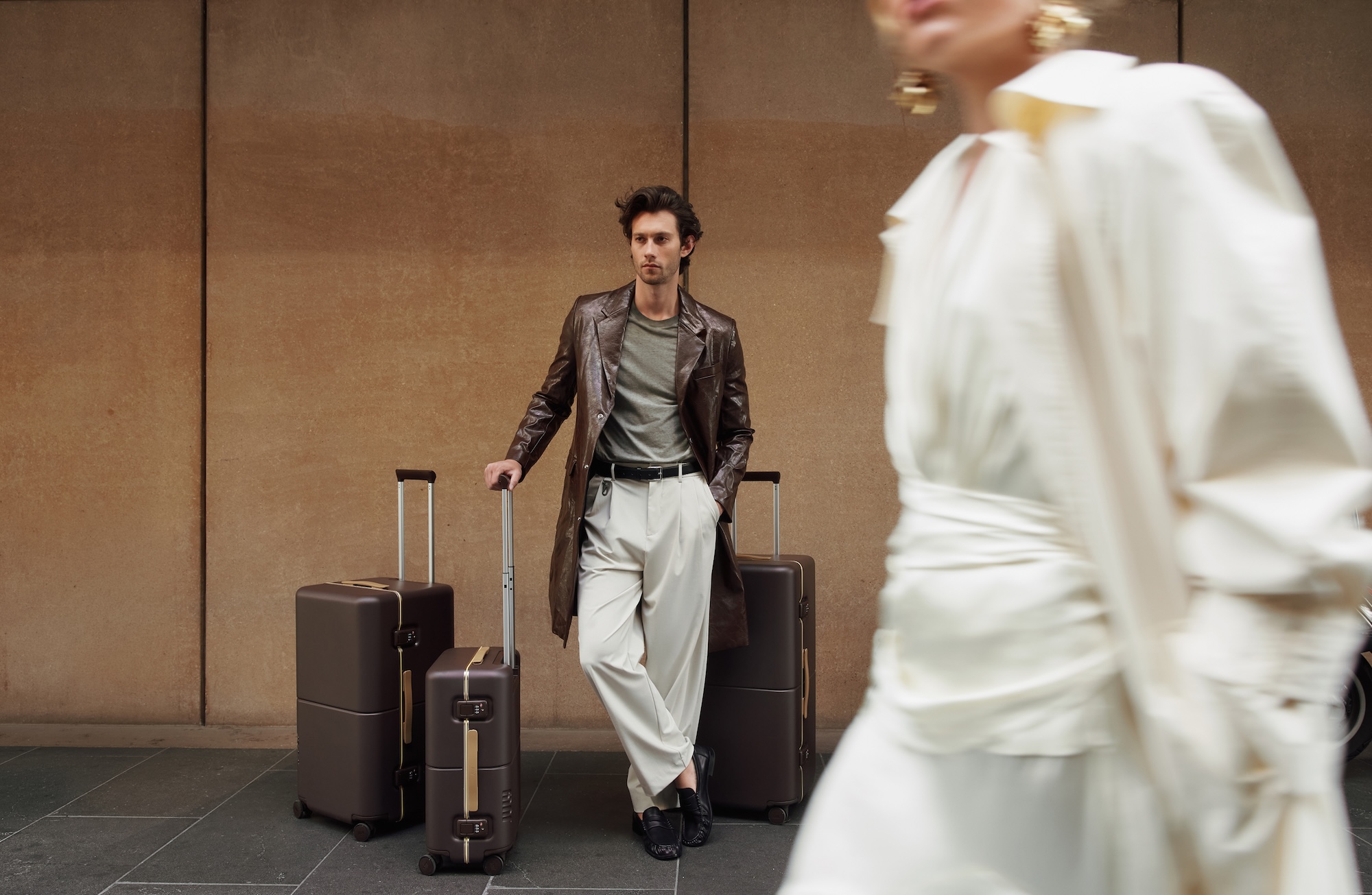 A man in a leather coat leans on July Trunk suitcases as a woman in cream walks past