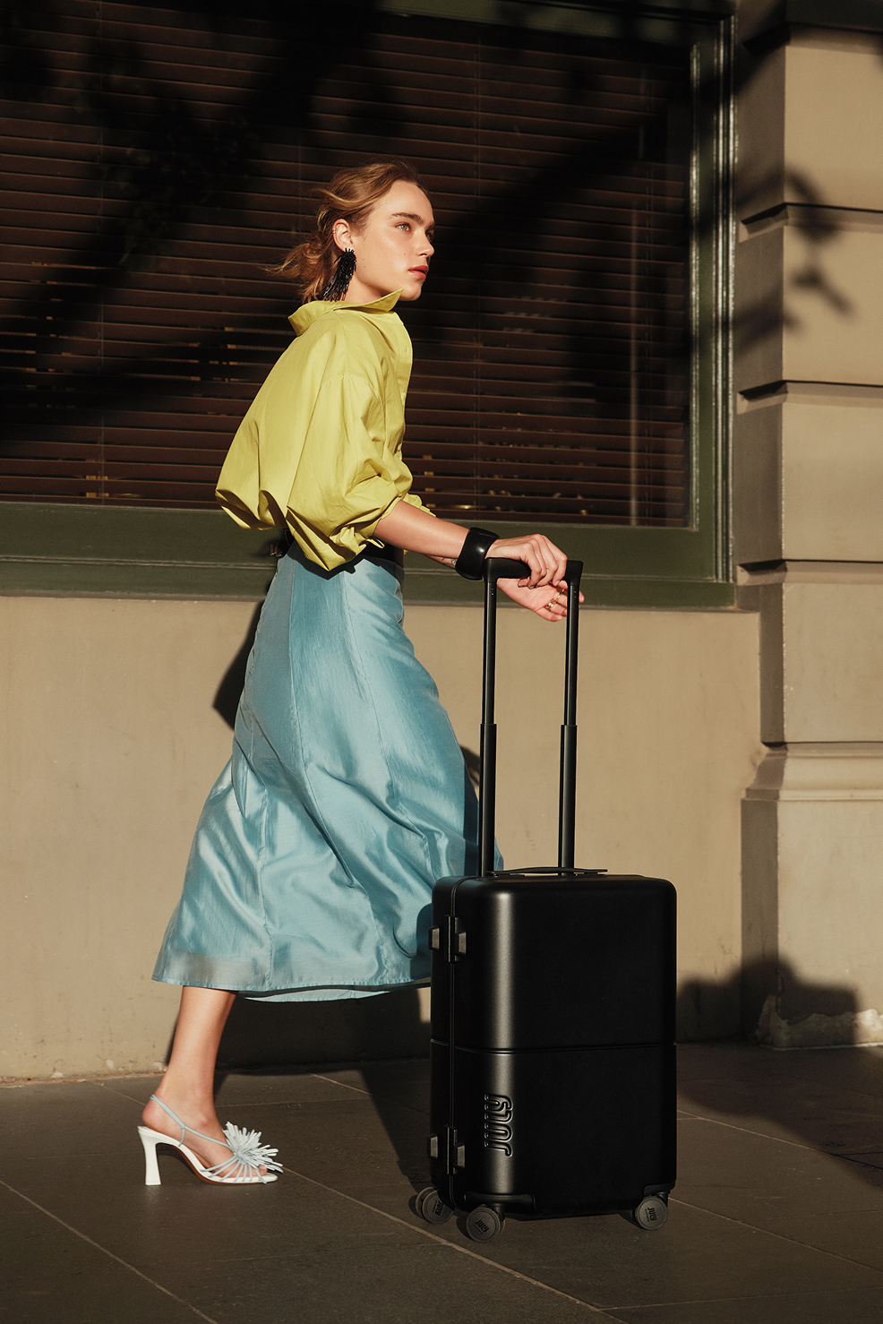 A woman in a yellow shirt and blue skirt wheels a black July Trunk down the street