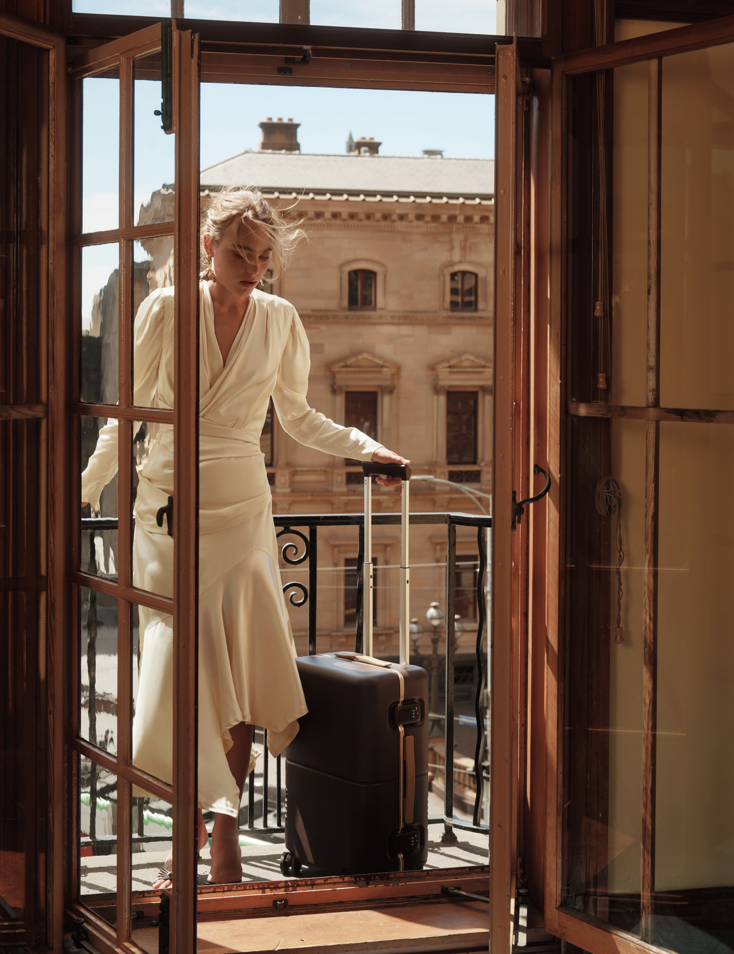 A woman in cream steps onto a balcony with a brown July Trunk